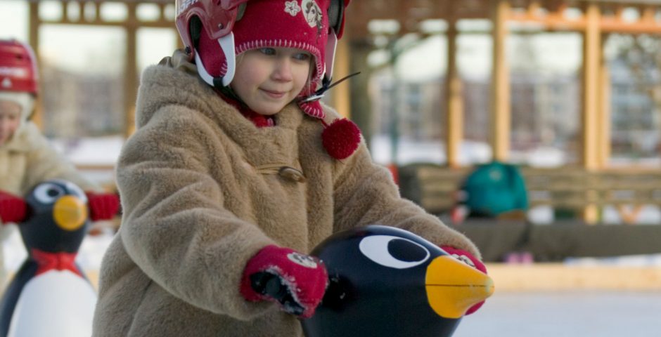 Barn med hjälm och vinterjacka åker skridskor tillsammans med Pingu.