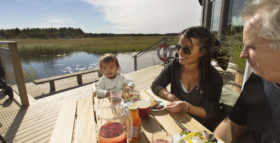 Morfar, dotter och litet barnbarn fikar tillsammans på bryggan till Naturum Värmland. I bakgrunden syns vattnet glittra, näckrosor och vassen på strandängarna.