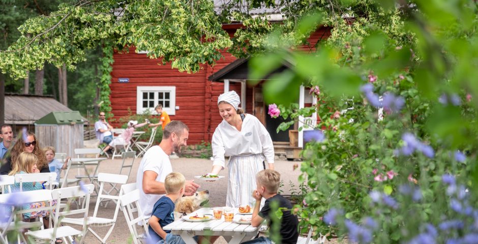 Kafépersonal serverar nygräddade våfflor till pappa med två söner, vid uteserveringen vid den röda timmerstugan Spikgården.